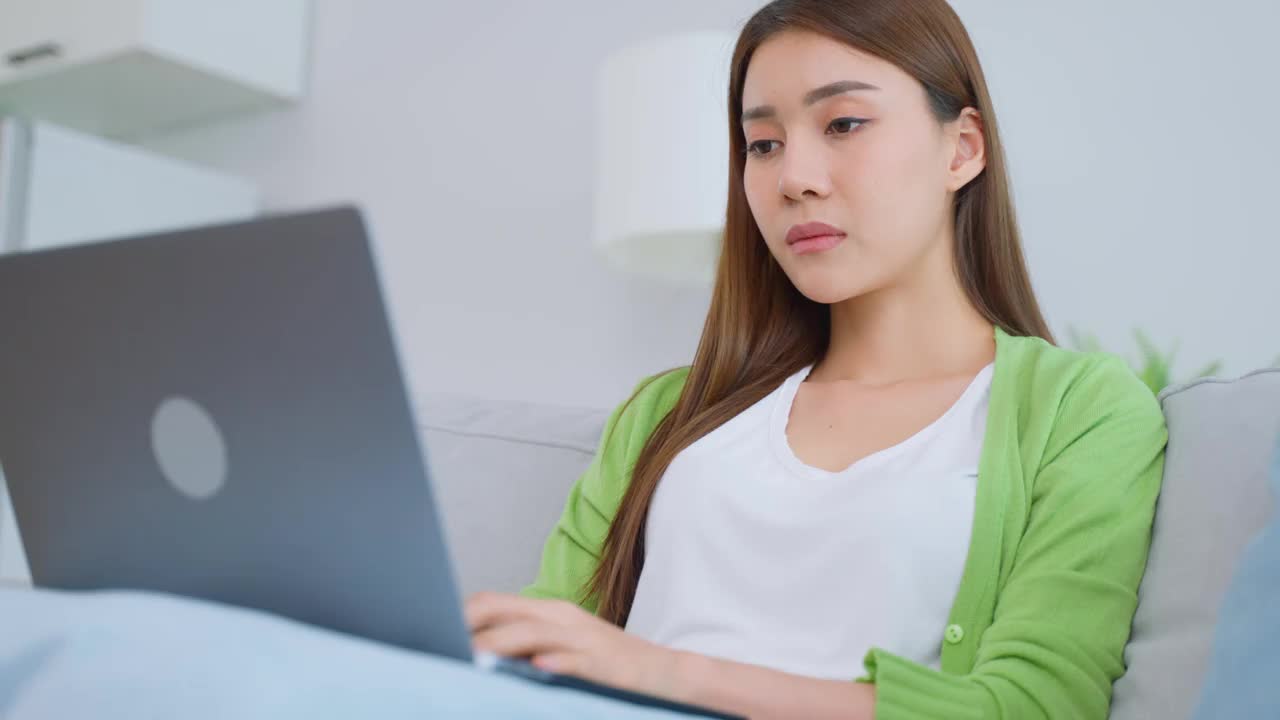 精疲力竭的亚洲女商人在使用笔记本电脑时伤了眼睛。年轻的女白领坐在沙发上感到视觉疲劳和眼睛疲劳，劳累劳累和按摩干燥易怒的眼睛在家里。视频素材