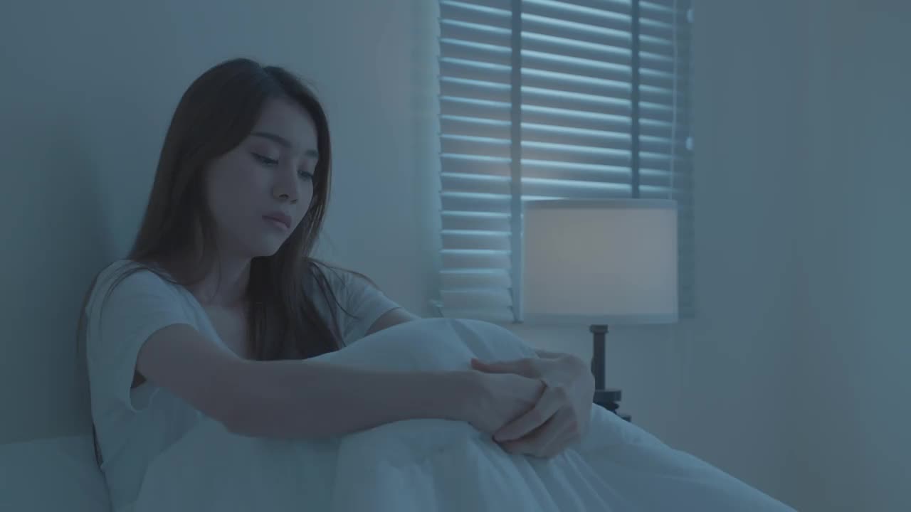 亚洲美丽沮丧沮丧的女孩独自坐在床上的卧室。有吸引力的不快乐的年轻女子感到悲伤，孤独和不安的生活问题，并试图伤害自己在黑暗的夜房在家里。视频素材
