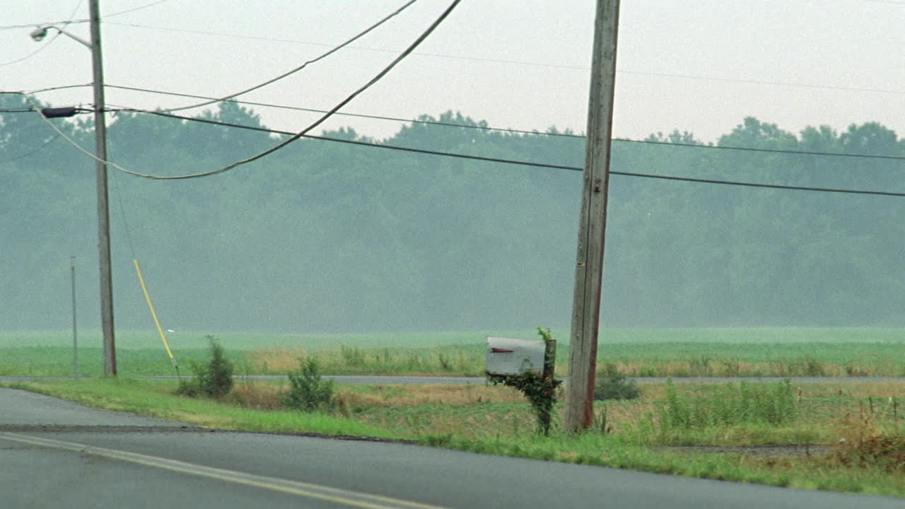 在一条双车道的乡村公路上，电话杆旁边是长满常春藤的广角信箱。在郊野可见的田野和树木、灌木丛或树林。天空是朦胧的。电话线在屏幕顶部可见。可能在农村或农村地区。视频下载