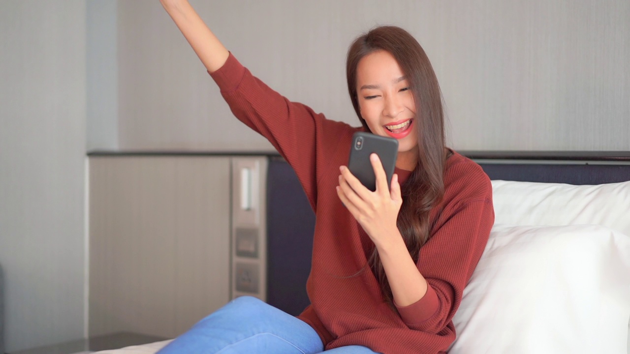 年轻的亚洲女性在床上使用智能手机视频下载