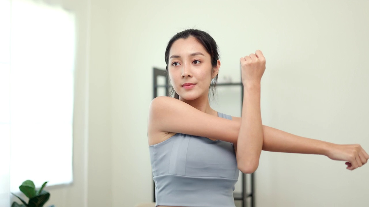 有吸引力的亚洲年轻健身女子伸展手臂热身之前在家里锻炼。美丽微笑的女性穿着运动服锻炼瑜伽在客厅。视频下载