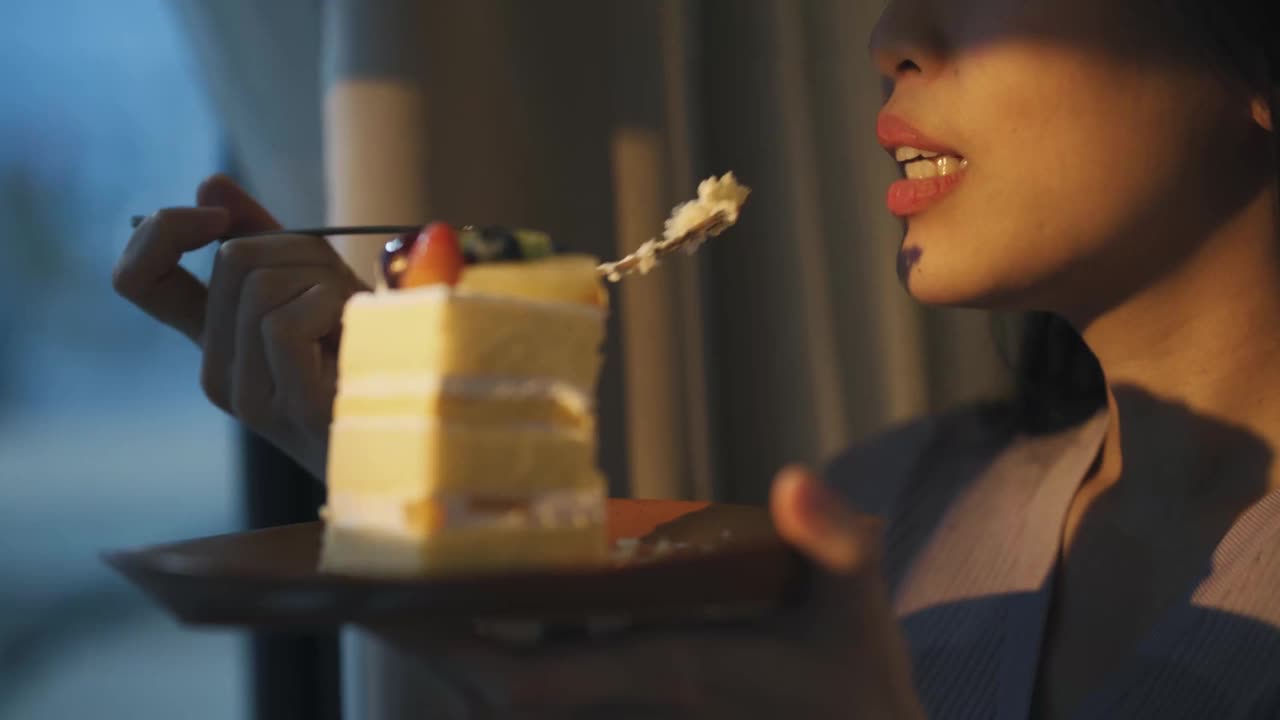 亚洲中国美女享受一片蛋糕在日落期间看着远方透过窗帘视频素材