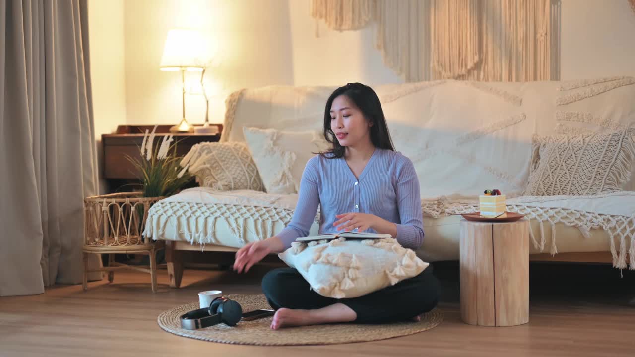 亚洲华人妇女享受她的空闲时间坐在客厅的地面上看书，听音乐，吃蛋糕在周末深夜视频素材