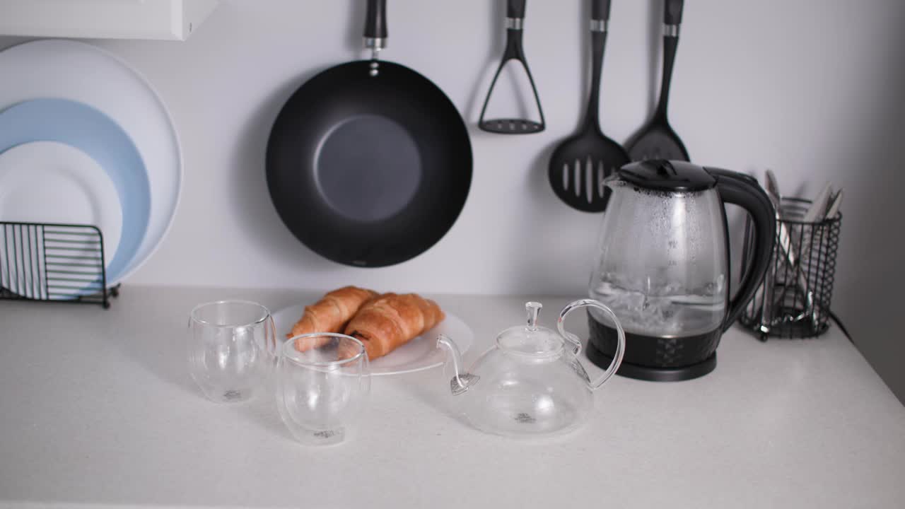 茶会上，一名妇女在厨房的杯子和羊角面包的背景上，把热水花草保健茶倒进玻璃茶壶里视频素材