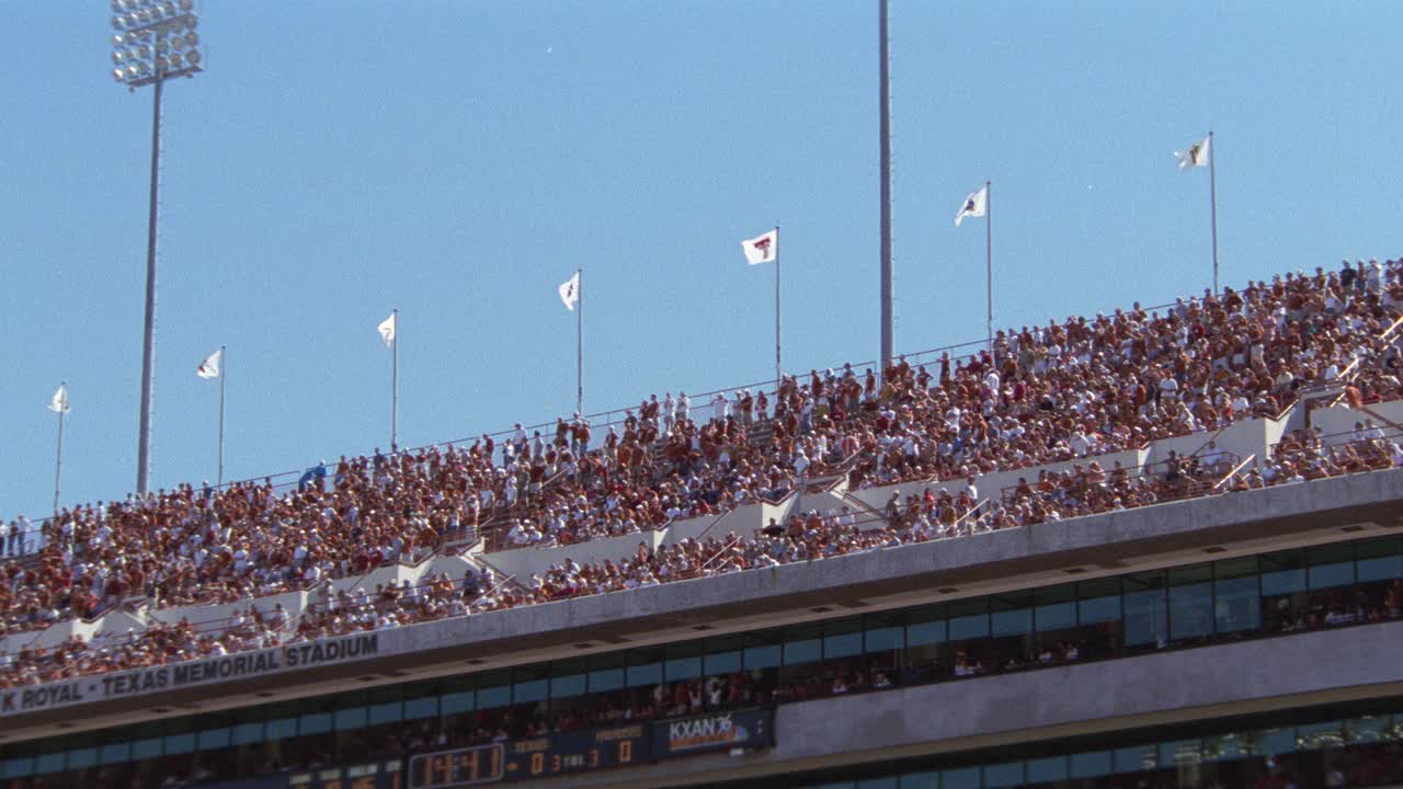 观看德克萨斯大学橄榄球比赛的观众或球迷在体育场欢呼的角度。达雷尔k.皇家纪念体育场。人群的欢呼声。观众上方的迷你记分板。大学橄榄球队的旗帜飘扬在环绕体育场顶部的柱子上。视频素材