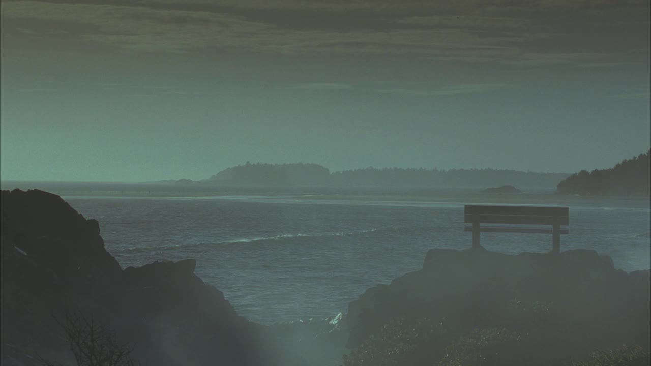 广角崎岖的海岸线与山脉和森林在bg。在fg中，面对大海的长凳坐落在一块大石头或圆石上。波。雾在空气中传播。视频素材