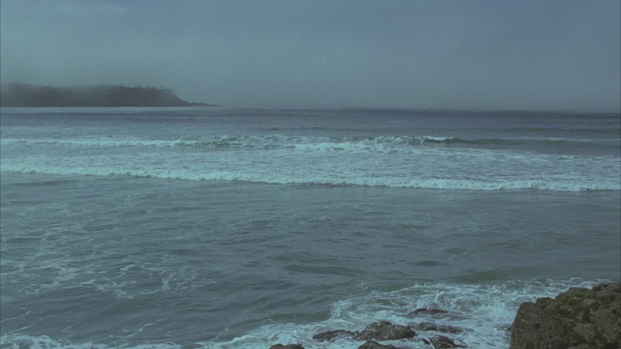 从右到左，从海浪冲击附近的岩石在海滩上到海滩上的房子或大厦崎岖的海岸线与山脉和森林在bg。雾。视频素材