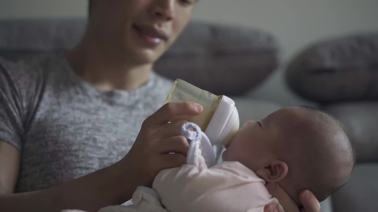 疲惫的亚洲中国年轻父亲打哈欠喂养他的儿子与奶瓶在客厅在周末视频下载