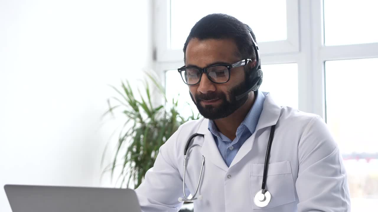 印度男医生穿着长袍，拿着听诊器坐在桌前，看着笔记本电脑视频素材