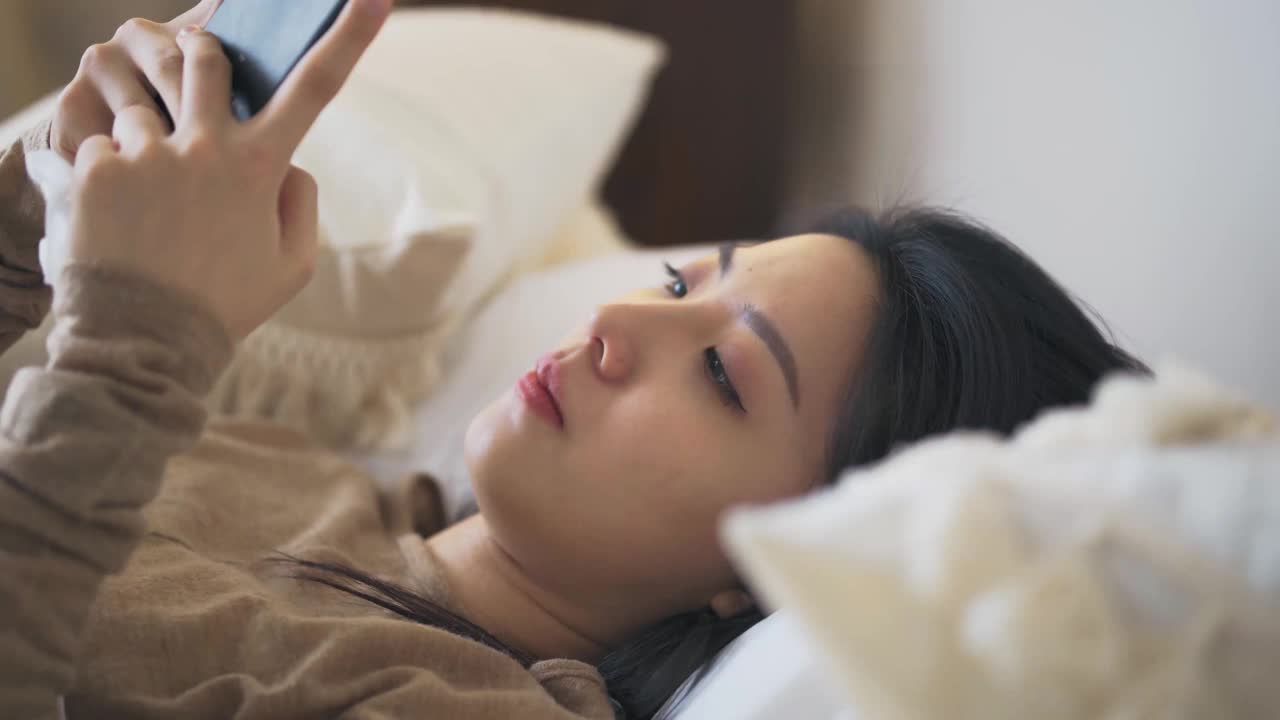一名亚洲华裔妇女躺在床上，盖着毯子，患感冒和流感，用智能手机和朋友聊天，用数字体温计测量体温视频素材