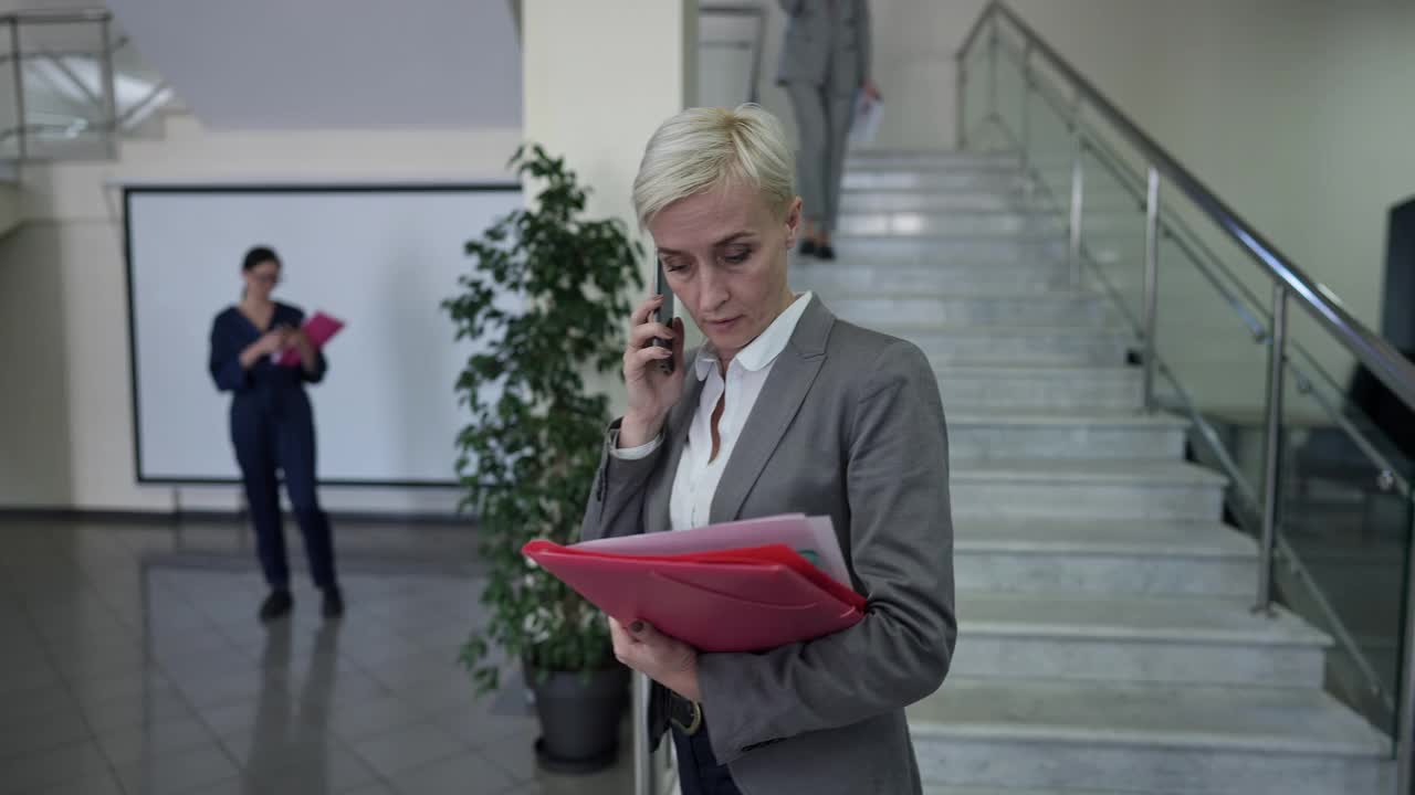 跟踪拍摄忙碌的成熟白人女性经理走在电话，路过同事在办公室大堂离开。成功自信美丽的女人在室内工作的肖像讨论的想法。视频素材