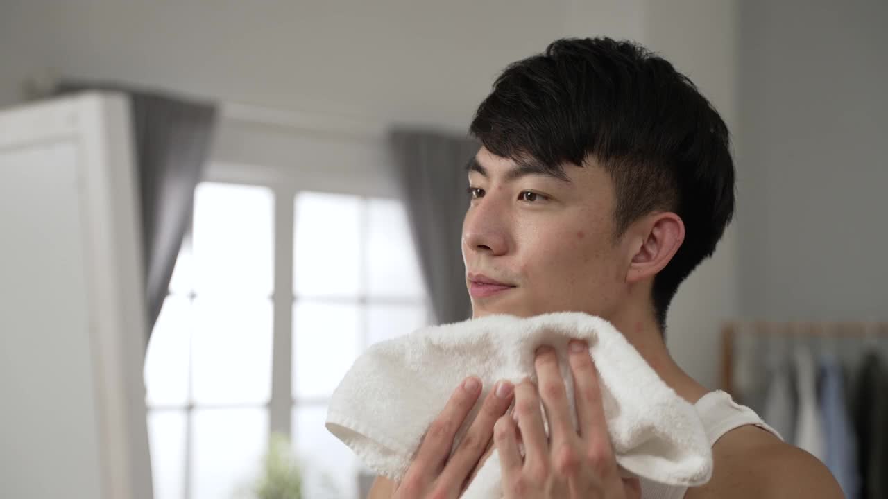 快乐的亚洲人穿着汗衫，看着镜子里的自己，用毛巾擦脸，每天早上在家里做皮肤护理视频下载
