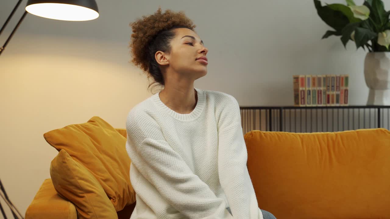 一名黑人年轻女子坐在客厅舒适的沙发上，闭着眼睛，双手放在脑后视频素材