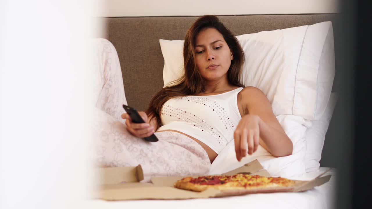 女人躺在床上吃披萨，一边用遥控器看电视视频素材