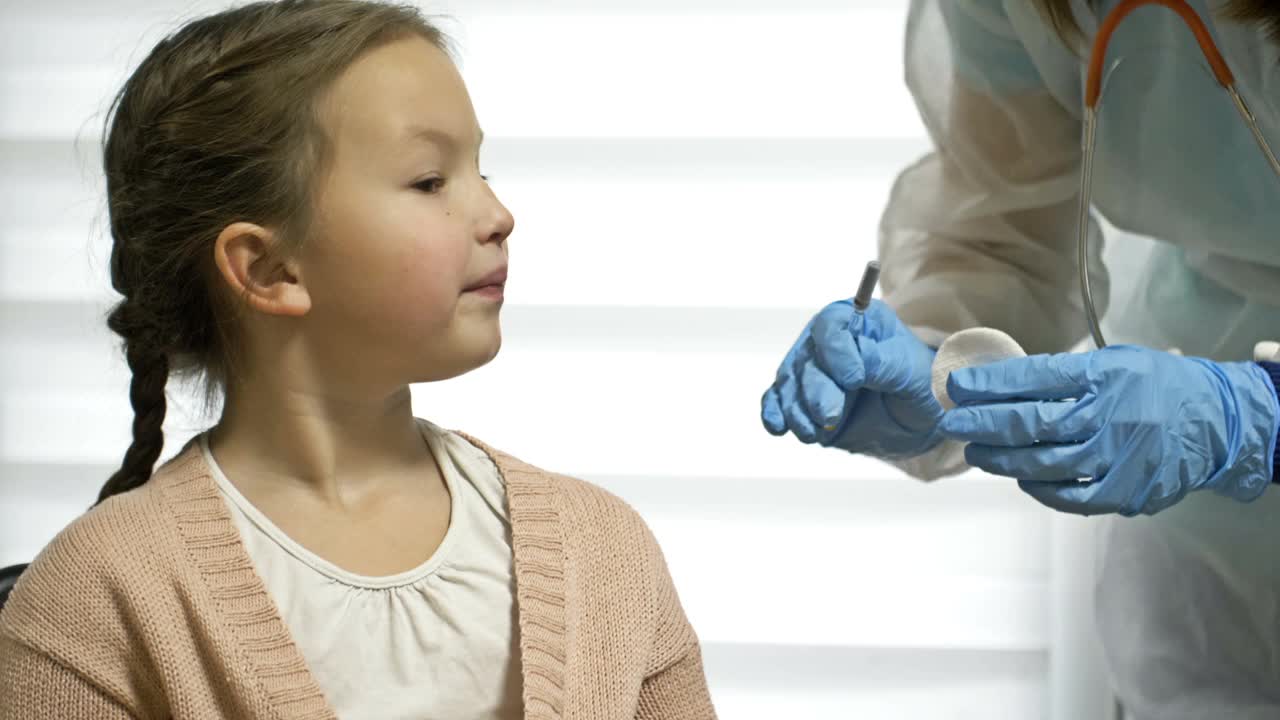 近距离拍摄的是穿着防护服、戴着医用口罩的专业医疗护理女护士，在实验室为可爱的小女孩注射新冠肺炎疫苗，接种概念视频素材