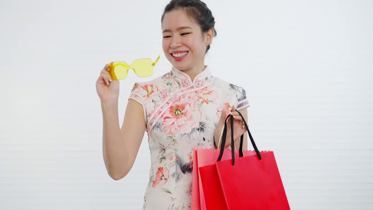 中国/亚洲妇女穿着旗袍，白色花卉图案的服装，在中国春节期间，黄色眼镜和购物袋，白色的背景视频下载
