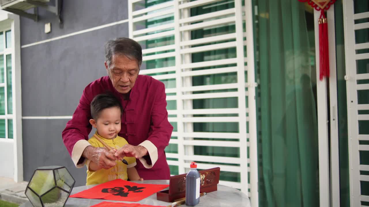 亚洲华人微笑的爷爷和孙子在中国新年期间在前院手写中国书法视频下载