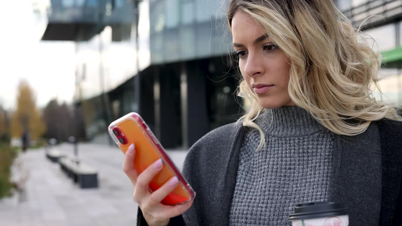 年轻漂亮的女商人喝着咖啡在办公大楼前用手机发短信视频下载