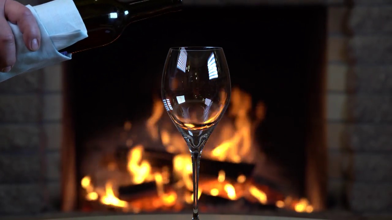 红酒倒进舒适的壁炉边的酒杯，燃烧的木头背景。视频下载