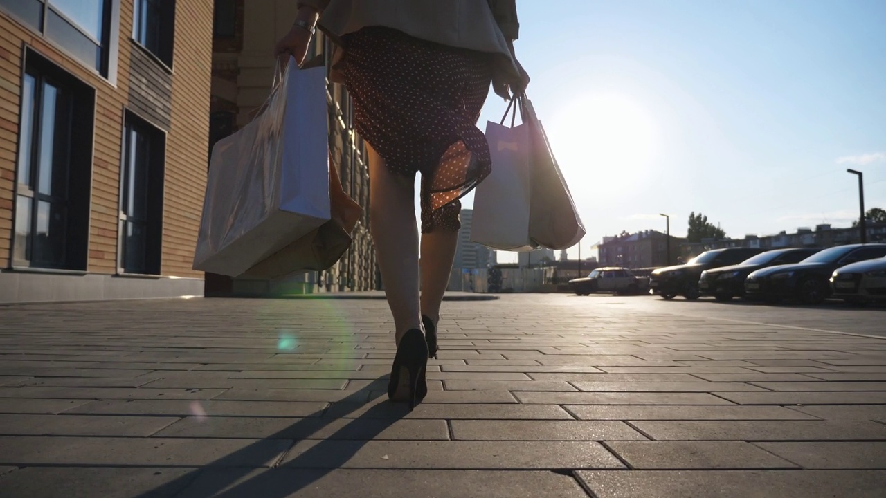 女性脚穿黑鞋，脚踩高跟鞋，走在人行道上。在城市广场买东西后拿着小包走的女孩。年轻女子的手提着购物袋走在城市的街道上。后视视频素材