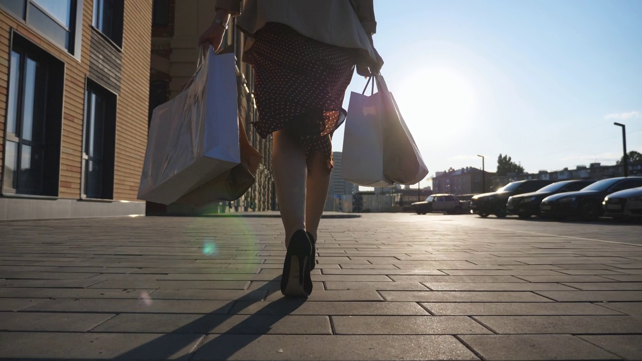 女性脚穿黑鞋，脚踩高跟鞋，走在人行道上。在城市广场买东西后拿着小包走的女孩。年轻女子的手提着购物袋走在城市的街道上。后视视频素材
