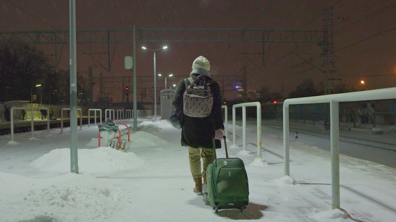 一个带着行李的成熟女人，一个游客，在冬天寒冷的雪夜走在火车站的月台上，等待着她的火车。视频下载