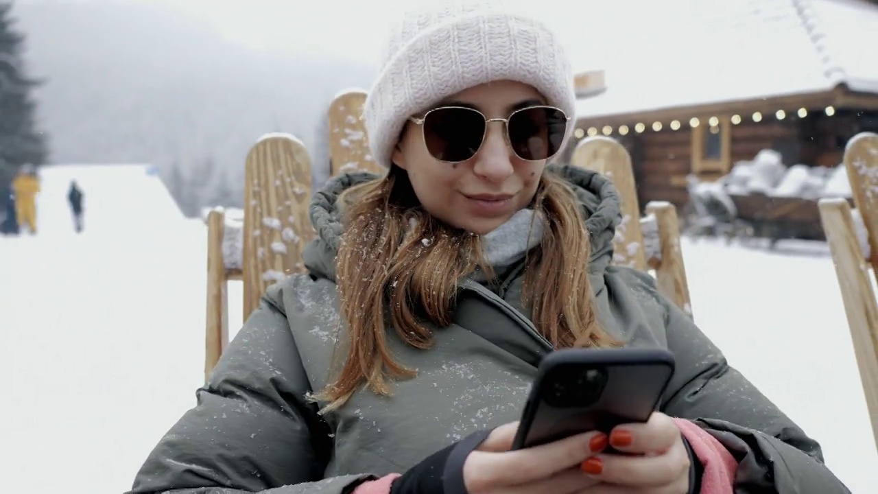一个时尚的年轻女孩在雪山下的一个度假胜地用手机自拍视频下载