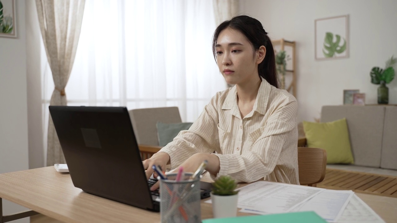 白天，在一间现代化的客厅里，穿着浅色衬衫、在家工作的漂亮的亚洲女商人正聚精会神地在电脑上打字视频素材