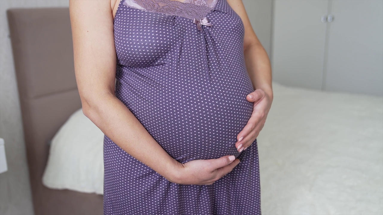 一个没脸的女人站在她的卧室里等着生孩子。怀孕最后一个月的女士抚摸着她的大肚子。准妈妈正在近距离地准备分娩。视频素材