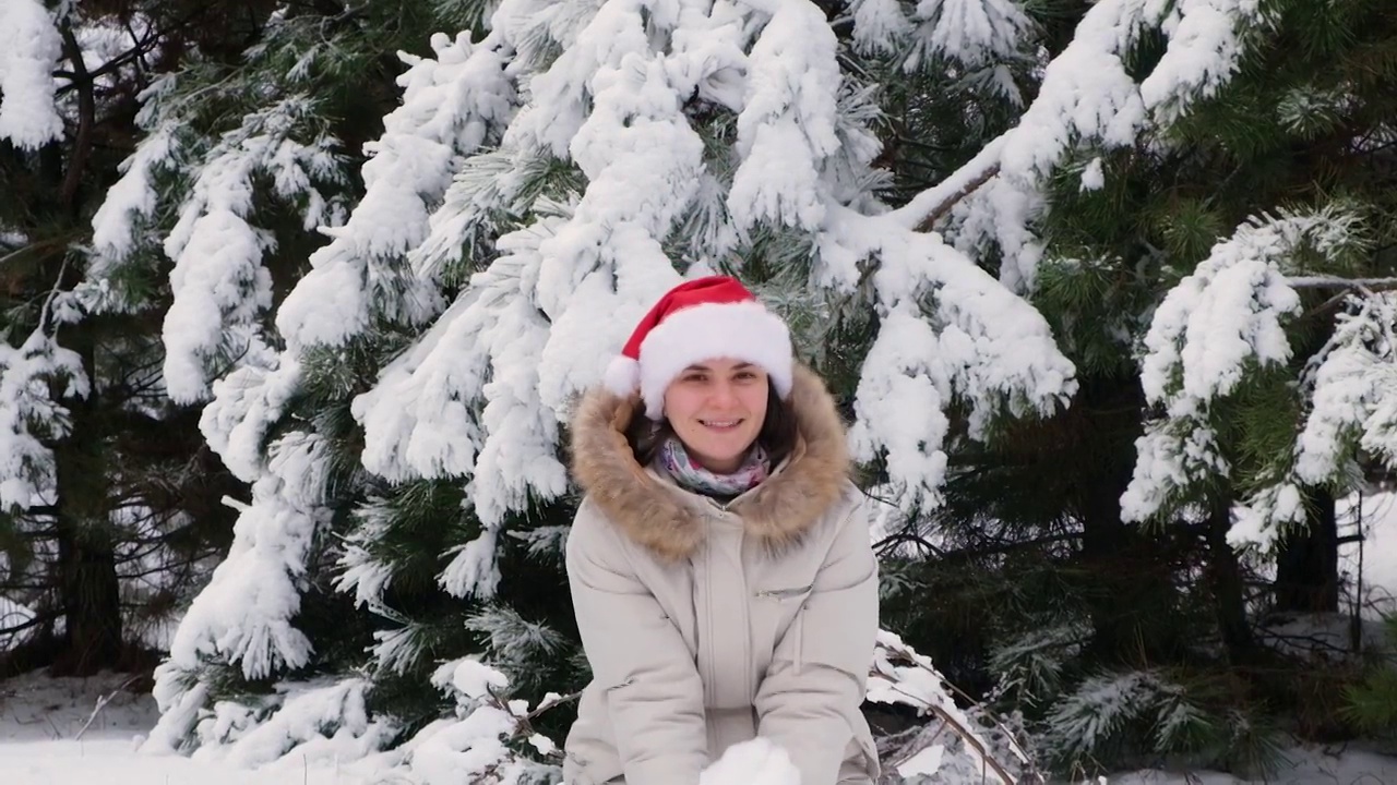 一个戴着圣诞帽的女人在冬天的松林里抛起雪花，圣诞节的心情。视频下载