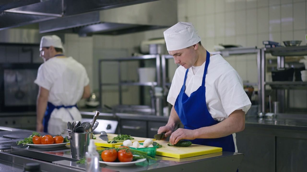 白种人厨师切黄瓜片吃片厨师路过背景。穿着制服的男人在餐厅室内的商业厨房工作的肖像。工作及职业观念。视频下载