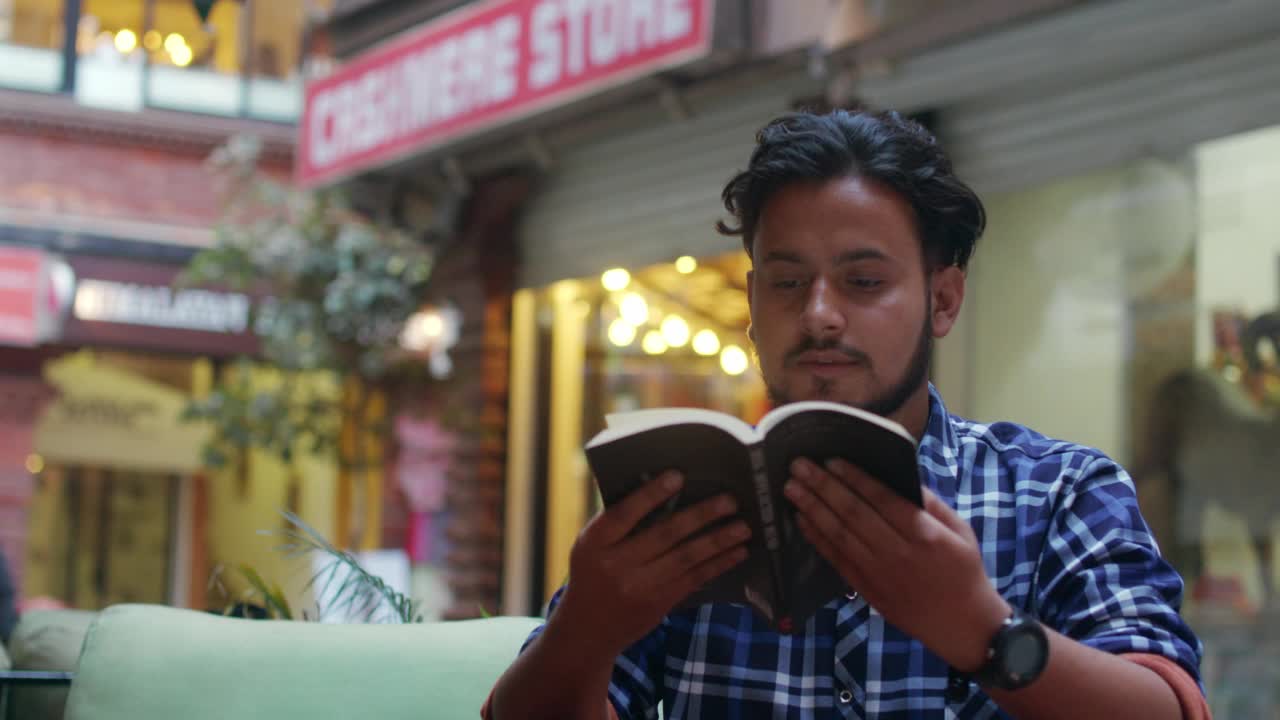 一位年轻的现代男性坐在户外的路边咖啡馆里全神贯注地看书。视频下载