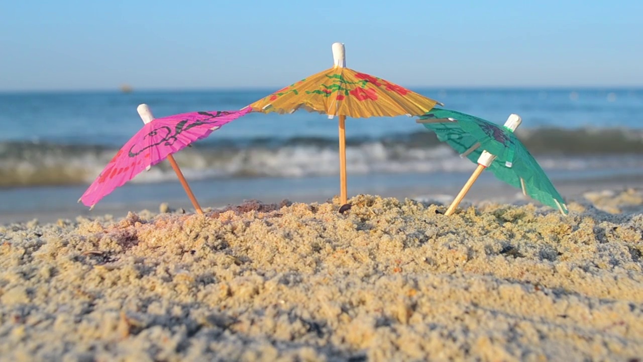 三把小纸鸡尾酒伞站在沙滩上的沙滩特写。沙滩上的小纸伞，在阳光明媚的夏天，海边的海浪。休闲，度假，旅游，旅游概念视频下载