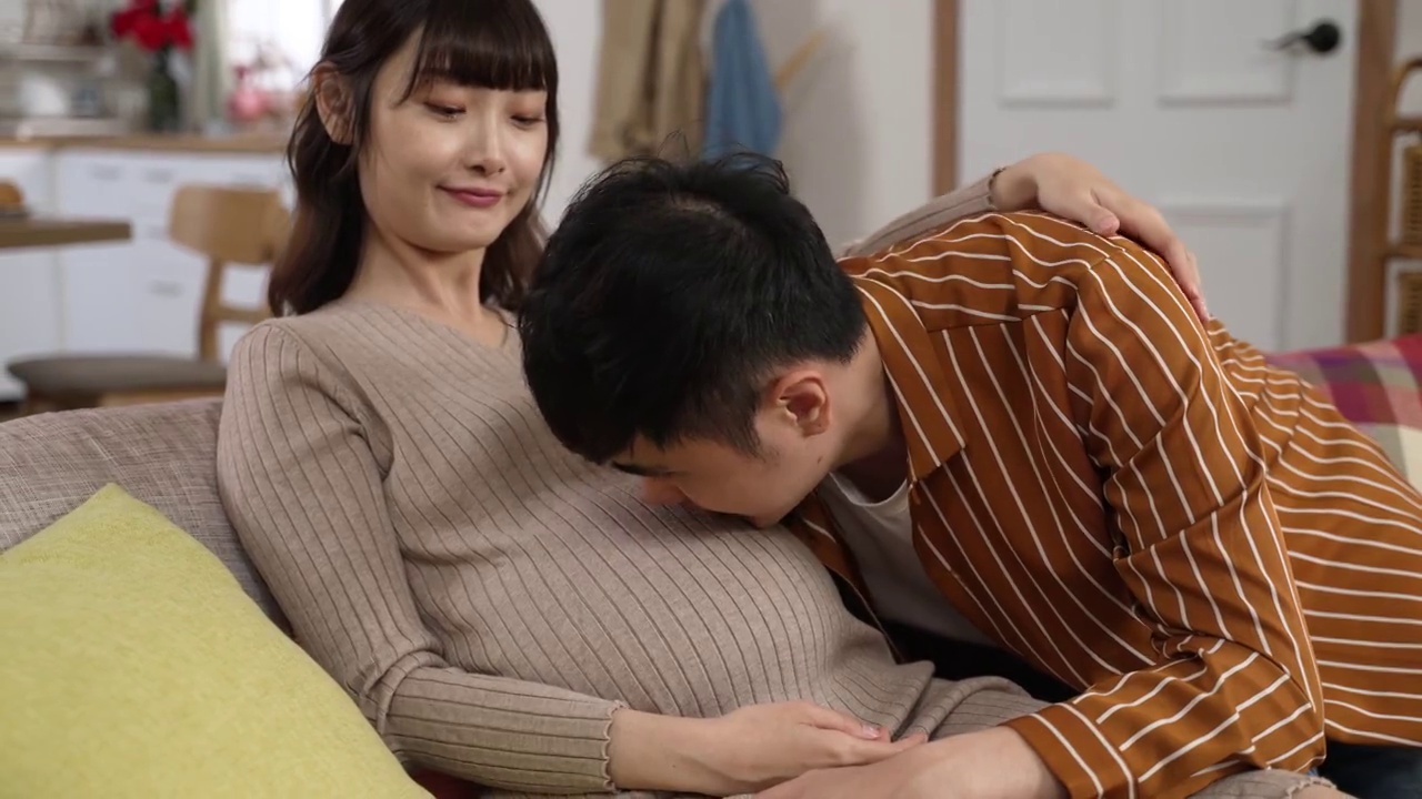 英俊的亚洲男人躺在他怀孕的妻子的肚子上说话，听胎儿，而他们在家里的客厅放松视频素材