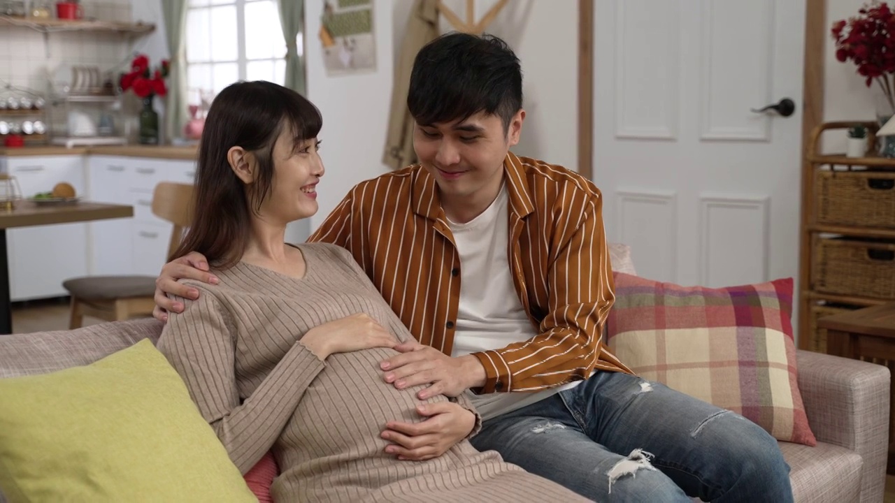 幸福的亚洲夫妇在家里客厅的沙发上放松时抚摸着怀孕的肚子。视频素材