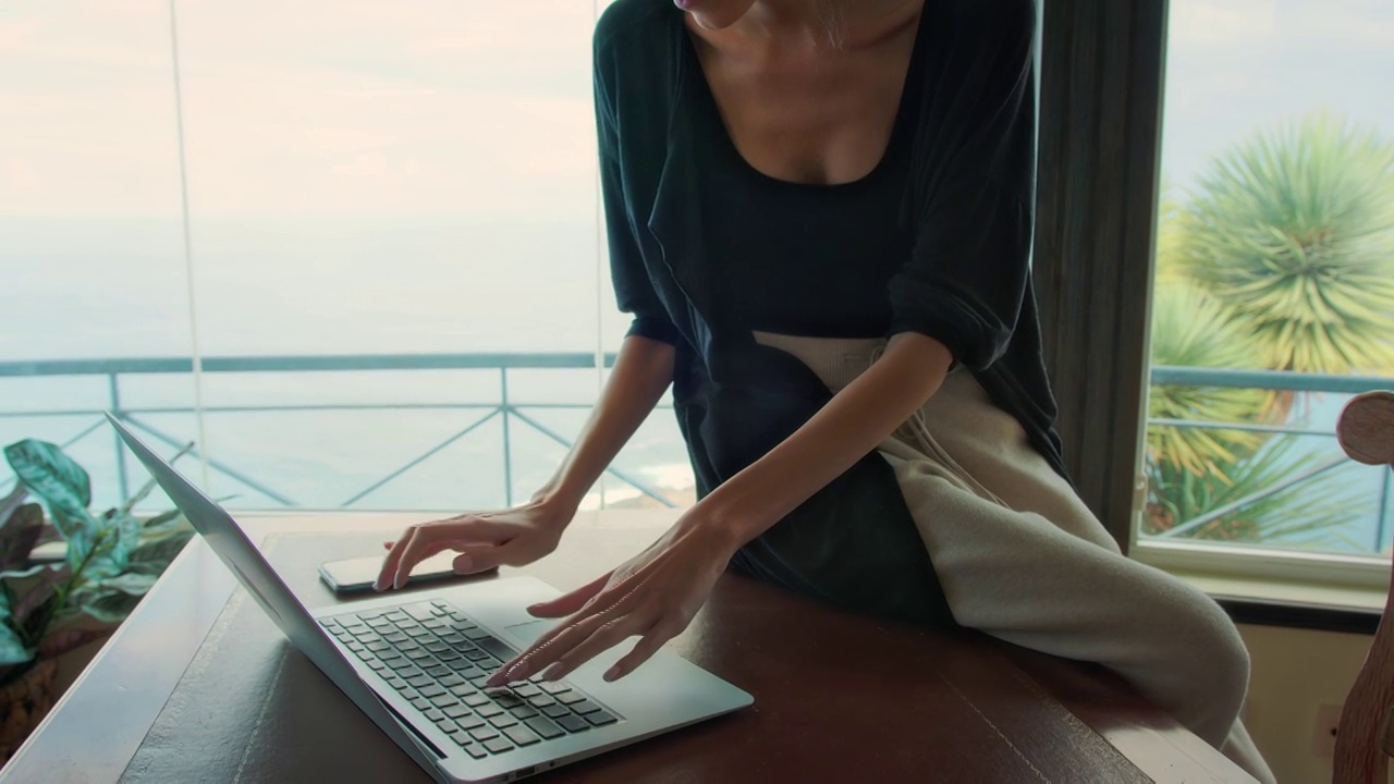 在全景窗旁工作的女人。使用笔记本电脑，喝咖啡，欣赏壮丽的海景视频下载