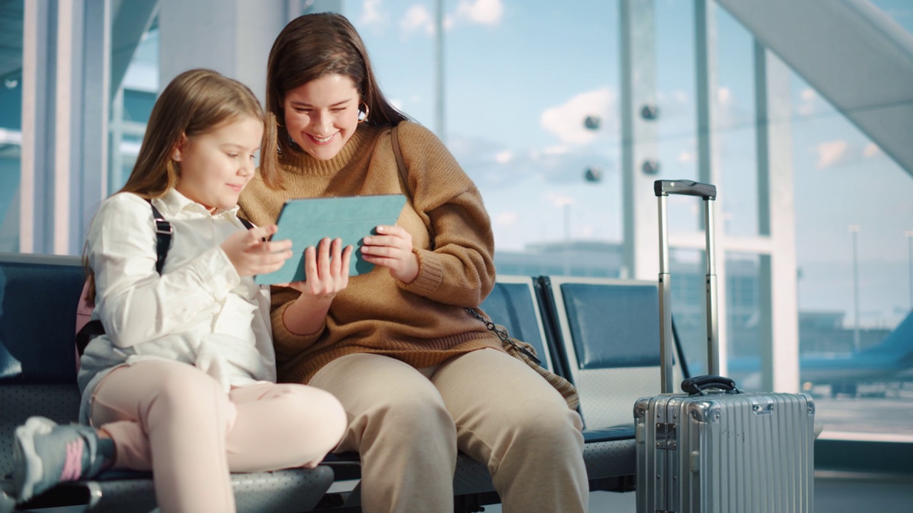 机场候机楼:可爱的妈妈和小女儿在等待他们的假期航班，在数码平板电脑上玩教育游戏。年轻的家庭坐在飞机飞行的航空枢纽的登机休息室视频下载