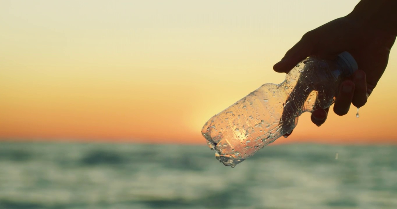 慢镜头的特写，一个年轻的志愿者正在从海上捡起一个塑料瓶，以保护夕阳下的环境。视频下载