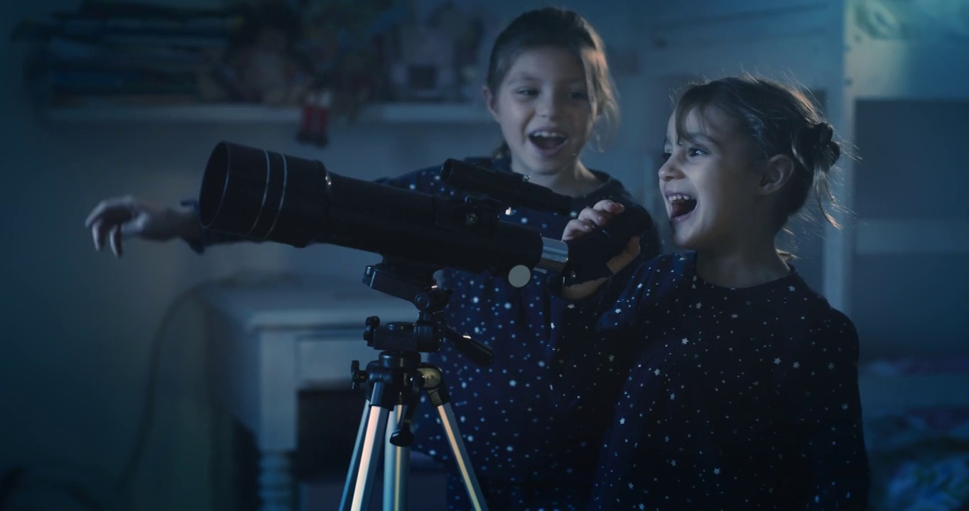 在夜晚拍摄的电影中，穿着缀满星星的睡衣的两个快乐的小女孩正在通过望远镜观看黑暗的天空中坠落的星星。概念:探索、教育、童年视频下载