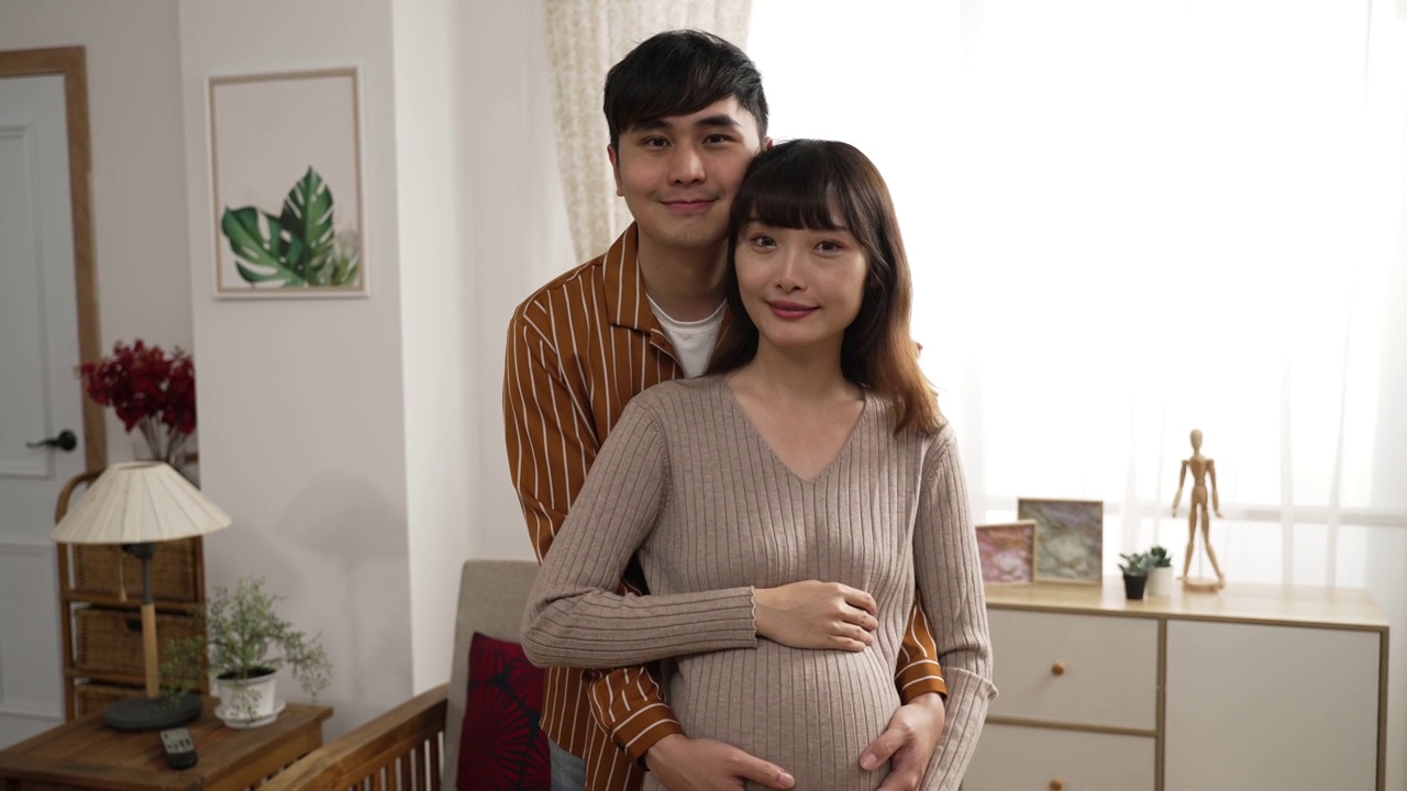 未来的父母站在家里看着相机微笑的半身像。丈夫从背后抱住妻子的肚子视频素材