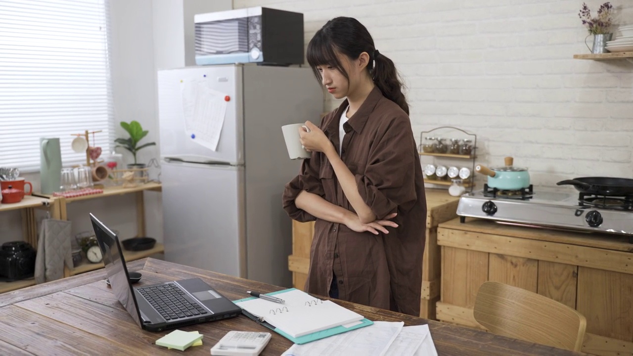 亚洲女性创意工作者一边喝着茶，一边沉思着看着电脑，一边在家里工作视频素材