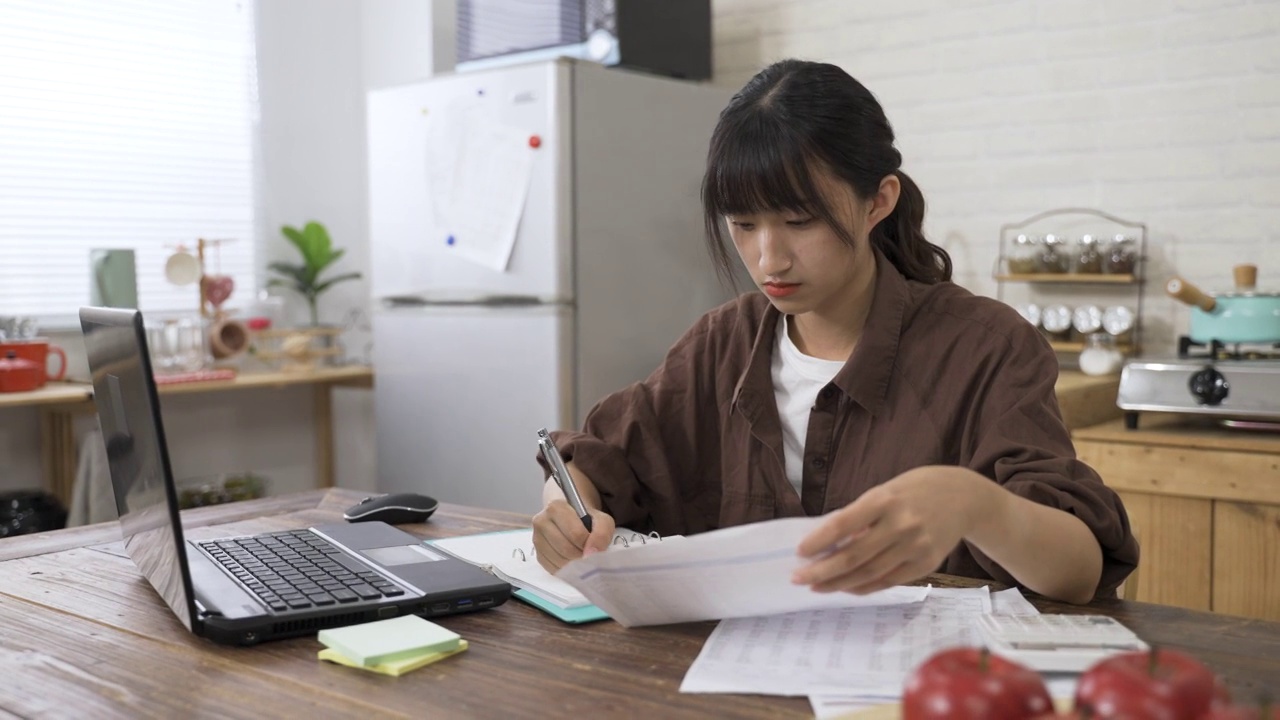 图为，白天在家里的餐厅里，一名集中精力的中国女性一边用笔记本电脑在线申报纳税，一边在笔记本上记录数据。视频素材