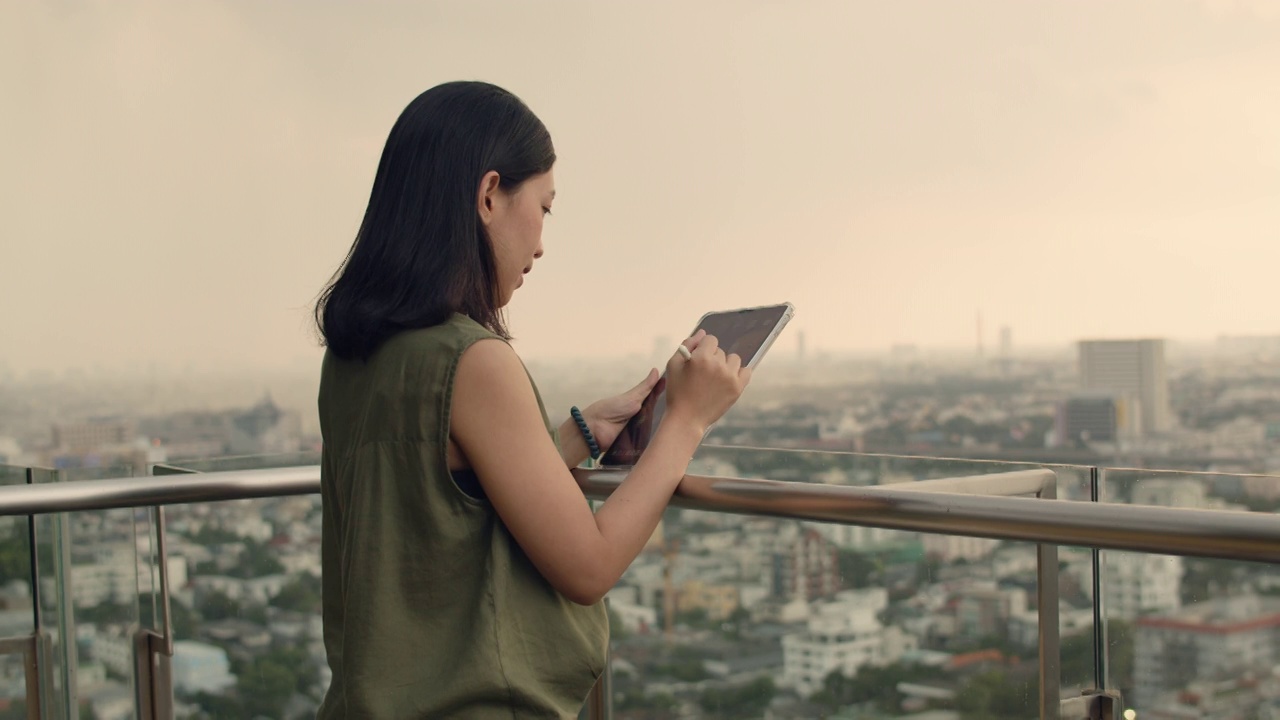 一名亚洲女性在日落时分站在公寓屋顶的阳台上用平板电脑和朋友聊天。视频素材