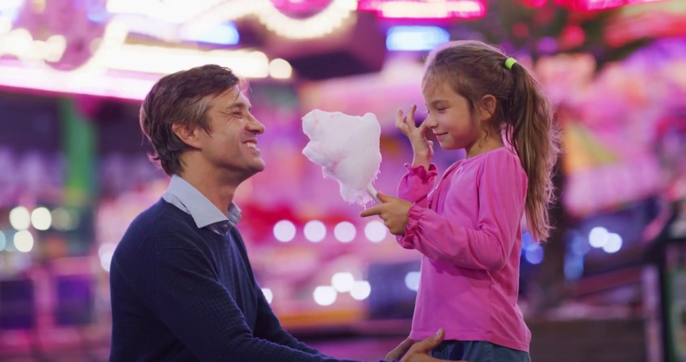 真实的照片，一个快乐的父亲和他的小女儿正在开心地吃棉花糖在游乐园与月亮公园的灯光在晚上。视频素材
