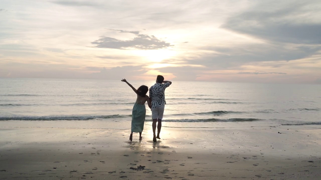 亚洲女性旅行者握着男人的手，看着美丽的日落在海滩上，夫妇在夏天度假和自由的概念视频购买