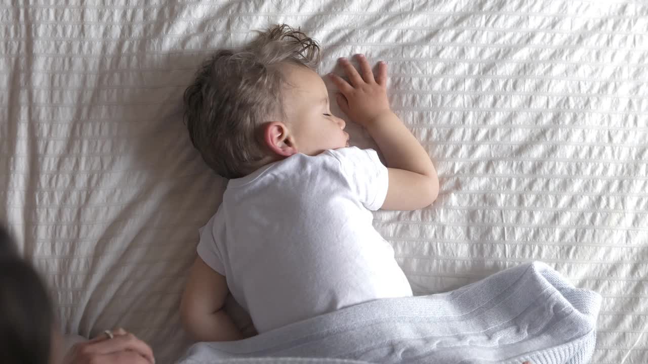 真实俯视图的母亲正在爱抚她的蹒跚学步的男婴，白天睡在床上柔软的毯子在托儿所。家庭的概念，舒适，关心，爱，甜蜜的梦，母亲，童年。视频下载