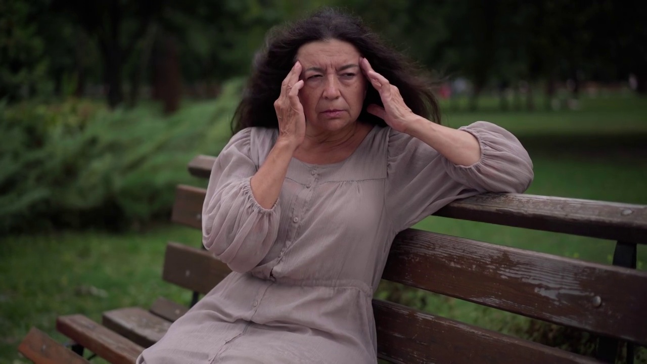 我累了，伤心的老女人擦太阳穴坐在公园的长凳上。白人退休人员与头痛偏头痛户外在风阴天的肖像。慢性疾病与生活方式概念。视频素材