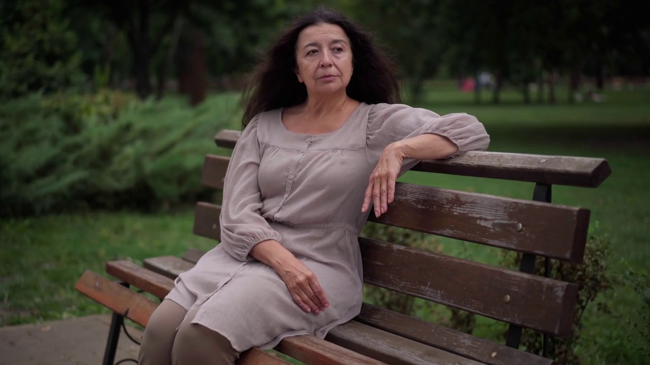 老年白人妇女坐在公园的长椅上突然头痛。在阴天多风的日子，患病的女性退休人员偏头痛户外的肖像。医药保健理念。视频素材
