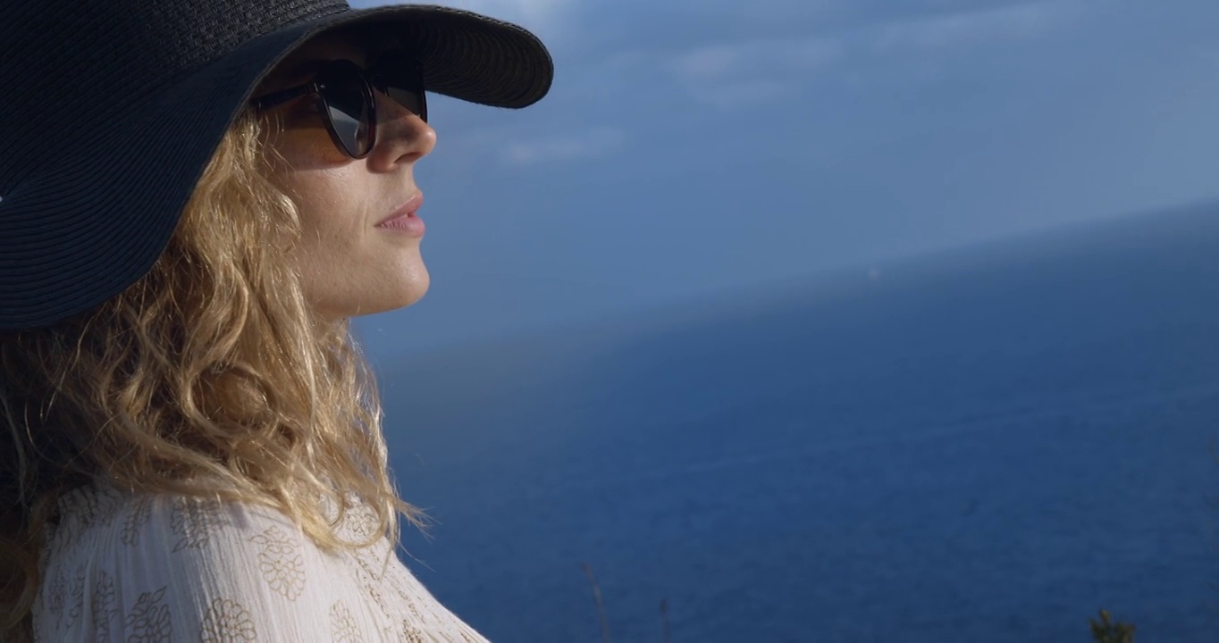 2剪辑!一个年轻美丽的女人凝视着大海，穿着飘逸的衣服，戴着一顶帽子。米诺卡-西班牙4K视频下载