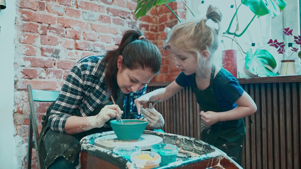 年轻的妈妈和她的小女儿在陶轮上学习陶艺，在现成的碗上画画。视频下载